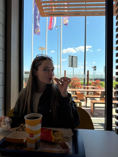 A young woman with long, light brown hair is eating a french fry in what appears to be a McDonald's restaurant. She is wearing a black jacket and has sunglasses perched on her head. In front of her is a tray with a McDonald's cup and a container of french fries. Outside the window, there are several flags, including what appears to be a Russian flag and McDonald's flags. The scene is likely in the city of Ub Municipality, Serbia, given the prompt context.