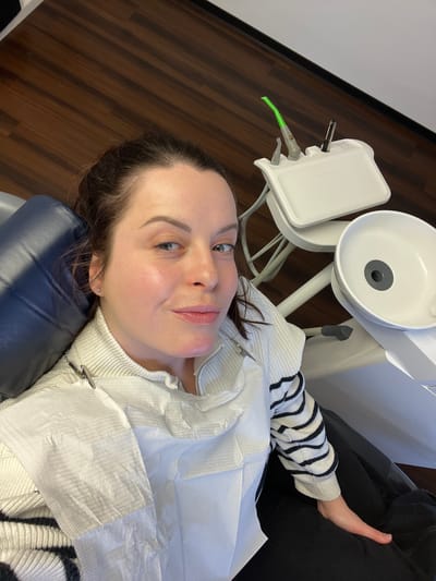 An overhead-angle photograph depicts a female subject seated in a dark blue dental chair within a dental office in Monsheim, Germany. The subject, a woman with brown hair pulled back, fair skin, and light-colored eyes, directly gazes at the camera with a slight smile. She is wearing a light-colored, possibly off-white, ribbed turtleneck sweater over a black and white striped long-sleeve shirt. A white, textured disposable dental bib is secured to her sweater at the neckline by a silver clip on a chain. To the subject's right, a white dental unit is visible, featuring a tiered instrument tray holding multiple dental tools, including one with a distinctive green tip, and a white ceramic spittoon with a gray drain. The floor is composed of dark brown wood laminate, and a white wall with a dark baseboard is visible in the background.