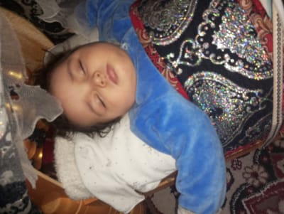 A baby is sleeping in a cradle. The baby is wearing a blue and white patterned garment. The baby is resting on a white fleece-like material, with a decorative pillow behind the baby's head. A sheer, lace-like fabric is partially covering the cradle. The cradle appears to be wooden. The scene is in Galaosiyo, Uzbekistan.