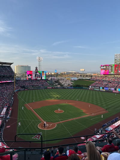 The image depicts an overhead view of Angel Stadium during a baseball game in Anaheim, California, United States. The baseball field is visible with players in position. The stadium is packed with spectators, and large screens display game information. The stadium is surrounded by city buildings. The sky is clear blue.