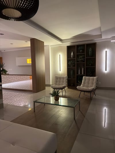 A contemporary living space features a dark brown shelving unit with decorative objects, flanked by two white cushioned chairs with black frames. A clear glass coffee table sits on hardwood flooring with a potted plant atop. The room is illuminated by linear LED lights. A wooden partition leads to an area with textured white wall panels and another plant. A black, circular decorative piece is visible in the foreground.