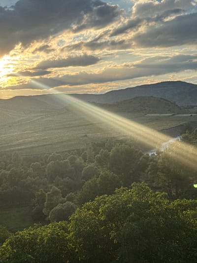 The image depicts a serene landscape in Aspindza, Georgia, bathed in the warm, golden light of the setting sun. The sky is a dramatic canvas of dark and light clouds, with distinct beams of sunlight piercing through, creating a radiant, ethereal glow that illuminates the scene. In the foreground, lush green trees with vibrant foliage dominate the lower portion of the frame, their leaves catching the sunlight and appearing almost luminous. Behind them, a gentle slope covered in a mosaic of green fields stretches into the middle ground. A winding river or stream is visible, reflecting the light and adding a touch of shimmer to the landscape. Further in the distance, rolling hills and mountains rise, their silhouettes softened by the fading daylight. The overall impression is one of tranquility and natural beauty, a picturesque moment captured at the end of a day in the Georgian countryside. There are no people or specific man-made structures clearly visible in this natural landscape scene.