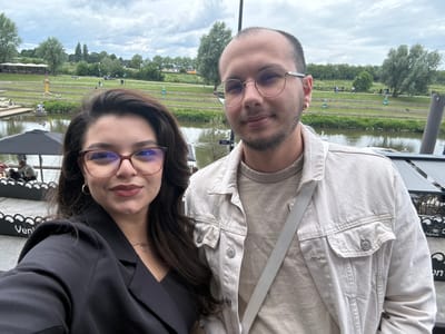 Two individuals, a woman and a man, are captured in a close-up selfie-style photograph. The woman, positioned on the left, has dark hair, wears reddish-framed glasses, a black blazer over a dark top, a gold hoop earring in her visible ear, and a delicate necklace. The man, on the right, has a shaved head, a beard, and wears round, gold-rimmed glasses, a light beige denim jacket over a matching t-shirt, a gold hoop earring in his left ear, and a light gray strap across his chest. Both individuals are looking towards the camera with slight smiles.
The background reveals an outdoor setting identified as Venlo, Netherlands. Directly behind the subjects is a body of water, likely the Maas River, with a partially obscured sign reading "Werf" visible on its edge. To the immediate left, a black decorative barrier displays the word "Venlo" in white lettering. The mid-ground features a waterside restaurant or cafe area with black umbrellas and outdoor seating. Beyond the water, a large terraced green embankment rises, covered in grass and numerous trees, some with light green foliage. Paved paths wind through the green area, populated by several people walking, some with dogs, and others on bicycles or scooters. Distant white tent-like structures with umbrellas are visible on the upper levels of the embankment. The sky is overcast with dense clouds.