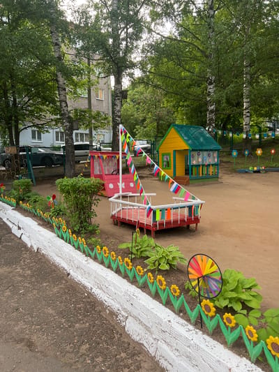 The image depicts a children's playground in Советско-Никольское, Russia. In the foreground, a white-painted concrete border separates a dark asphalt surface from a sandy playground area. This border is adorned with a decorative fence made of green zig-zag elements topped with yellow sunflower figures. Beyond the border, lush green plants, including hostas, are visible. A colorful pinwheel spins in the gentle breeze.

Further into the playground, a red and white structure resembling a boat is decorated with a string of triangular flags in red, white, and blue. Behind this, a small, brightly colored playhouse with a yellow exterior and a green roof stands under the shade of birch trees. A pink play vehicle with a steering wheel is positioned to the left of the playhouse. In the background, a multi-story building with light-colored brickwork and numerous windows suggests a residential area. The surrounding environment is characterized by mature trees with dense green foliage, indicating a summer season. The ground is primarily sand, typical of many playgrounds. No people are visible in the image.