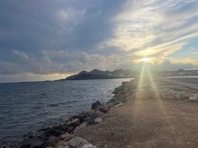 The image depicts a coastal scene in Cabo de Palos, Spain, captured during either sunrise or sunset. The left half of the frame is dominated by a body of dark blue-grey water, exhibiting minor wave activity. A rugged embankment of various-sized grey and brown rocks forms a jagged barrier between the water and a dirt/gravel pathway that extends along the right side of the image. Beyond this pathway, a lighter-colored, possibly concrete, pier or walkway structure is visible. In the mid-ground, a cluster of white and light-colored buildings belonging to a town or village is situated along the distant coastline, backed by undulating, dark green-grey hills or mountains. The sun is positioned low on the horizon, slightly right of center, emitting a prominent, bright white and yellow lens flare that emanates upwards and downwards from its source, partially obscuring portions of the sky and distant land features. The sky above is partly cloudy, with darker, heavier clouds on the left transitioning to brighter, more scattered clouds on the right, illuminated by the sun's glow. No individuals or discernible activities are present in the scene.