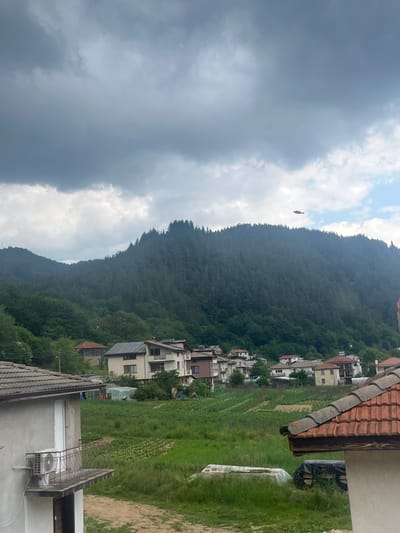 The image captures a village scene in Elhovets, Bulgaria, set against a backdrop of densely forested mountains under a cloudy sky. The upper left portion of the sky features dark, heavy grey clouds, gradually lightening towards the right where patches of blue sky are visible. A dark, fish-shaped object, possibly a kite, is suspended in the air in the upper right quadrant, above the mountain range.

The background is dominated by steep mountains covered in a dense expanse of dark green coniferous trees. In the mid-ground, several residential structures are visible. A prominent, light-colored, multi-story building with a roof featuring numerous solar panels is situated towards the mid-left. Other houses in the distance exhibit traditional red and grey tiled roofs.

The foreground on the left displays the corner of a light grey building with a white door, a small balcony featuring an ornate metal railing, and an exterior air conditioning unit mounted below the balcony. This building has a grey tiled roof. On the right, the corner of another building with a distinctive red-tiled roof is partially visible. Between the foreground and mid-ground structures, an expanse of green agricultural land with visible rows of cultivation is present, traversed by an unpaved path. No individuals or animals are discernible in the scene.