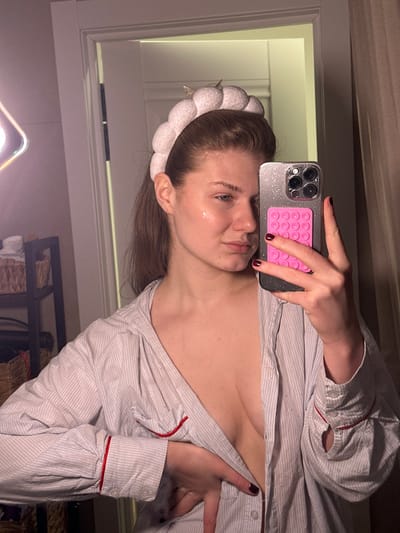 The image displays a mirror selfie of a female individual, observed indoors. The described location is the city of Yanga-Yul, Russia.
The subject, a female with light brown hair, wears a white, textured headband with a subtle golden accent. A reflective sheen is present on her left cheek. She is dressed in a light-colored, vertically striped button-down shirt, unbuttoned to expose her upper chest. The shirt features long sleeves with red piping visible on the cuffs. Her fingernails are painted with dark maroon polish. She holds a smartphone, identifiable by its multi-lens camera array and a glitter-finish case, in her right hand. A pink, multi-suction cup accessory is affixed to the phone's back. Her left hand is positioned over her midsection, partially obscuring the shirt's buttons. Her expression is neutral as she looks towards the mirror.
The reflected environment includes a white door frame behind the subject. To the far left, a square-shaped, illuminated LED panel is partially visible. Adjacent to the light, a dark-colored, multi-tiered shelving unit or basket stand is partially visible, containing woven baskets. A light-colored curtain or wall is present on the far right.