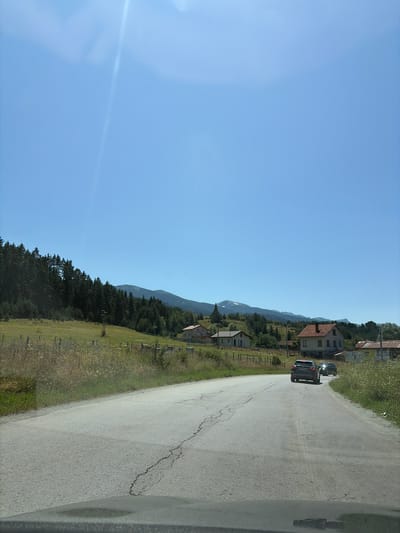 A wide shot captures a sunny day in Madzhare, Bulgaria, with a clear blue sky dominating the upper half of the frame. The lower half of the image depicts a paved road with visible cracks, curving slightly to the right as it recedes into the distance. Two dark-colored cars are driving on the road, one behind the other.

To the left of the road, a grassy field stretches out, bordered by a dry stone wall and sparse vegetation. Further back, a dense pine forest covers the lower slopes of rolling hills. In the distance, the silhouette of a mountain range under a hazy sky is visible.

On the right side of the road, several houses with red-tiled roofs are scattered across the landscape. The houses appear to be residential, with some having gardens or yards. The environment suggests a rural or semi-rural setting.
