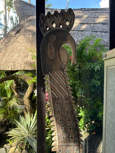 The image displays a vertically oriented, elaborately carved wooden totem or sculpture in the city of Tibubiyu, Indonesia, positioned in what appears to be a doorway or framed opening. The sculpture, dark brown with lighter carved details, features a stylized bird-like head at the top, characterized by an elongated, downturned beak and a five-pointed crown with internal cutouts. Its elongated body has a sawtooth-patterned left edge and intricate carvings across its surface: diagonal stripes in the upper section and multiple rows of repeating S-shaped scroll patterns enclosed in diamond/square motifs in the lower section.
The background reveals a vibrant outdoor tropical environment. To the left of the sculpture, a large, conical thatched roof structure is partially visible, indicating traditional Indonesian architecture, beneath a clear blue sky with visible palm trees. Lush green foliage, including variegated succulent plants and other tropical trees, grows near the base of the roof structure. To the right of the sculpture, dense green bushes and trees with small red blossoms are visible, alongside a light-colored wooden door or panel with a dark hinge. The ground cover in the outdoor areas consists of dark soil, rocks, and small stones. The scene is brightly illuminated by natural daylight. No individuals are present in the image.