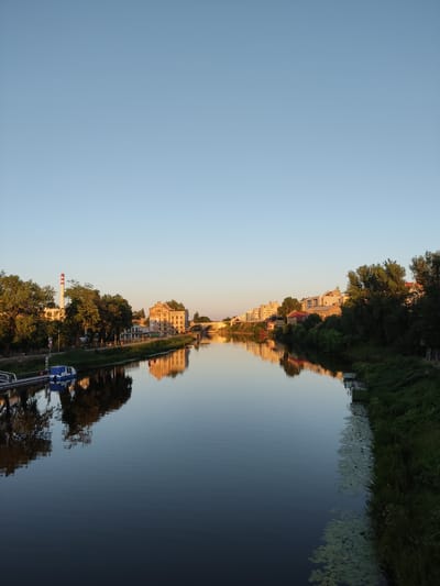 A wide-angle, eye-level shot captures a tranquil river scene in Kolín, Czechia, during what appears to be late afternoon or early evening. The clear, pale blue sky dominates the upper half of the frame, with a gentle gradient towards the horizon. The Elbe River flows calmly, its surface acting as a near-perfect mirror, reflecting the buildings and trees lining its banks.

On the left bank, a tall, red and white striped industrial chimney stands prominently, accompanied by several lush green trees. Adjacent to these, a small blue and white boat is docked. A signpost with a red circle and a diagonal line, indicating no entry or prohibition, is visible near the boat. Further along this bank, a series of buildings, some with ornate architecture, are visible, their reflections shimmering in the water.

The right bank is densely covered with trees and vegetation, with more buildings peeking through the foliage in the distance. A concrete structure, possibly part of a dock or embankment, is present at the water's edge, with water lilies floating nearby. A bridge, featuring multiple arches, spans the river in the middle distance, connecting the two sides of the city. The overall impression is one of serene urban riverside life, bathed in the soft light of a setting or rising sun, creating a symmetrical and peaceful atmosphere.