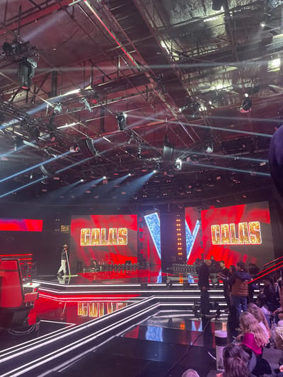 A tilted view captures a stage setup with a colossal lighting rig above, featuring numerous spotlights and metal framework. Two large screens display "CHILLS" in golden letters on a red background, flanking a blue starry display. On the stage, performers and audience members are visible, their figures partially obscured. White stripes create a visual effect leading towards the stage, complementing the vibrant colors of the set.
