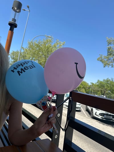 An outdoor scene, under a clear blue sky, features two balloons held by a person. The left balloon is light blue and has "Happy Meal" printed on it in black. The right balloon is pink and has a black smiley face drawn on it. The person's arm and blonde hair are visible on the left side of the frame. They are holding the balloons with their right hand, which has nails painted with a black and white spotted design. A pair of black-rimmed glasses are hanging on a dark railing in the foreground. In the background, trees and a street with white cars are visible. The location is the city of Húmera-somosaguas-prado Del Rey, Spain.