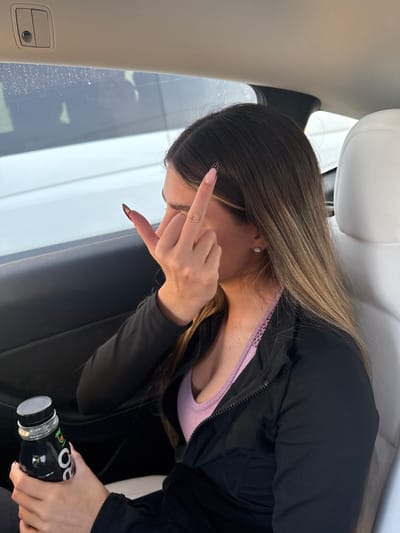A female subject is depicted seated within the passenger compartment of a vehicle, in Rosh Haayin, Israel. The subject, seen from the mid-chest upwards, has long, brown hair with lighter ends. She wears a black, partially unzipped athletic jacket over a light purple top, and a silver stud earring in her right ear. Her right hand is raised, performing a gesture where the index and pinky fingers are extended, and the middle and ring fingers are curled, commonly known as "devil horns" or "rock and roll" sign. Her fingernails are long, pointed, and feature a nude base with black, patterned tips. Her left hand holds a dark-colored plastic bottle with a black cap, displaying an orange logo. The vehicle interior shows a white headrest, black door trim, and a window reflecting a blurred white exterior.
