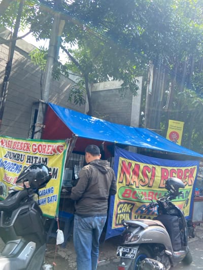 The scene is a street food stall in Kecamatan Cimahi Selatan, Indonesia, located at coordinates -6.909 latitude and 107.568 longitude. The stall is set up under a blue tarp awning. A man in a dark jacket and blue jeans is standing at the stall, likely ordering food. Behind him is a black scooter with a black helmet on the seat, and in front of him is a white scooter with a black bag on the back. The stall has a yellow and red sign advertising "Nasi Bebek Goreng" (Fried Duck Rice) with a black sign that says "Bumbu Hitam" (Black Seasoning) and a green and yellow sign that says "Nasi Berek" (Rice with a Side Dish). There is a red sign on a nearby post that says "Promo" and "Harga Terus Tanpa Kenaikan" (Prices Remain Unchanged). The stall appears to be operating in a residential area, as a concrete wall with a tree is visible behind the stall. The ground is a mix of gravel and pavement. The scene appears to be a typical street food stall in an Indonesian city, likely set up for the lunch rush.