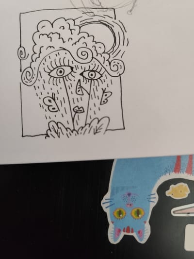 The image depicts a hand-drawn illustration of a stylized face with large eyes and a cloud-like hairstyle, set against a background of stylized grass and butterflies. The drawing is framed by a black rectangle. It is drawn in black ink on white paper. The drawing is placed on a black surface with the bottom of the page partially obscured by a blue, red, and white cat sticker. The cat sticker is looking towards the viewer with its eyes open, and it is partially obscuring a yellow fish sticker. The drawing is located on a surface in Clermont-Ferrand, France, at the coordinates 45.788 latitude and 3.11 longitude.