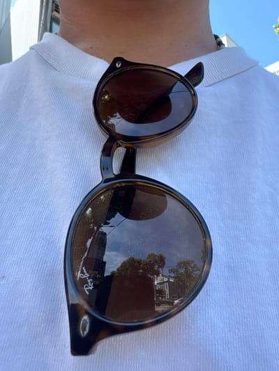 A pair of tortoise-shell Ray-Ban RB 2180 sunglasses rests on the white, ribbed polo shirt collar of an unidentified individual. A beaded necklace, featuring light blue/green and brown beads, is partially visible on the individual's right neck. The brown-tinted lenses of the sunglasses reflect an urban environment under a bright blue sky with some clouds. Reflected elements include a tall, dark modern building, a lighter-colored multi-story building, green trees, and a street light pole, indicating an outdoor setting in Jongno-gu, South Korea. The "Ray-Ban" logo is visible on the lower left lens, and faint, distorted Korean text consistent with local signage is reflected on the left lens, further confirming the location. Visible dust particles and smudges are present on the sunglass lenses.
