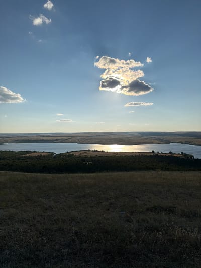 The image depicts a panoramic view of a landscape in the city of Gornyi Balyklei, Russia. The scene is dominated by a large body of water, likely a lake or reservoir, reflecting the bright sun. The sky above is a clear blue, punctuated by a few scattered clouds, one of which is illuminated by the sun, creating a halo effect.
In the foreground, there is a dry, grassy hill. Beyond the water, rolling hills stretch into the distance, covered with sparse vegetation and fields. A line of dark green trees runs along the edge of the water, separating the water from the drier landscape. The overall impression is one of a vast, natural setting under a bright, sunny sky. The time of day appears to be late afternoon or early evening, given the angle of the sun and the long shadows. There are no people or man-made structures clearly visible in the image.