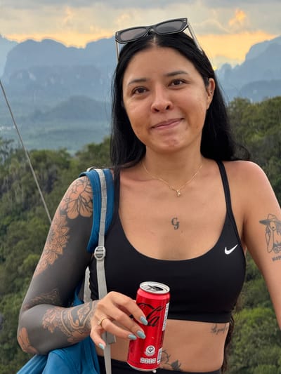 A woman with dark hair and tattoos on her arms and chest stands in front of a mountain range with a sunset sky. She is wearing a black sports bra and sunglasses on her head. She is holding a can of Coca-Cola in her right hand. The background shows lush green trees and misty mountains, characteristic of the landscape in Ban Khao Tang, Thailand. The lighting suggests it is late afternoon or early evening.