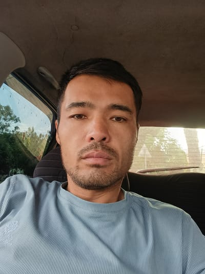 A man, likely of Central Asian descent, is depicted in a close-up selfie or portrait, seated inside a car. He has short dark hair, dark eyes, and a neat short beard and mustache. His expression is neutral and calm as he looks directly into the camera. He is wearing a light blue, short-sleeved top with a distinct ribbed texture. A subtle, light-colored embroidered design or emblem is visible on the left chest area of his shirt, though no specific text can be discerned.
The setting is the interior of a vehicle, with parts of the car's beige headliner visible above him and a dark seat or headrest behind him. Through the car's windows, an outdoor environment is visible, indicating it is daytime. The left window shows lush green trees and a bright sky, with what appears to be string lights or utility wires hanging against the horizon. The rear window on the right reveals more green foliage, a bright sky possibly indicating direct sunlight or haze, and a light-colored, triangular road sign (possibly a warning sign) partially visible through the glass. A rear windshield wiper blade is also seen at the bottom of the back window. The windows appear slightly dirty or dusty. The scene suggests the man is traveling or resting in a car during daylight hours, possibly in the morning or late afternoon, in Uchqo'rg'on, Uzbekistan. No other visible text is present in the image.