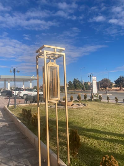 The image depicts an urban outdoor scene in Lkheng, Morocco, under a bright blue sky with scattered white clouds. In the immediate foreground, a tiled grey sidewalk runs along the left, adjacent to a strip of low, evergreen bushes. Dominating the foreground is a tall, golden-colored, geometric art installation or decorative lantern structure, composed of vertical and horizontal rectangular frames, encasing a central rectangular element with a reflective surface.

To the left of this structure, in the mid-ground, is a gas station with a white canopy bearing a green "V" logo. A white SUV with red markings and the Arabic word "إسعاف" (Ambulance) is parked beneath the canopy, with other vehicles partially visible behind it. Several dark outdoor chairs and tables are positioned near the gas station. Two black streetlights with double lamps are also present.

Further into the mid-ground, a well-maintained green lawn spans the area between the foreground and a multi-lane asphalt road. Within this lawn, a circular stone planter or fountain structure is visible, along with a line of small, conical evergreen shrubs. On the right side of the road, a tall, white vertical sign displays the red "TOYOTA" logo, with additional text beneath it. The background features scattered trees and bushes, along with low-rise buildings with reddish-brown roofs. No individuals are clearly visible in the scene.