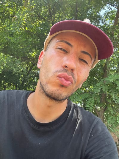 A man wearing a black t-shirt and a burgundy baseball cap is captured in a close-up selfie. His lips are puckered as if for a kiss, and he is looking towards the camera. The background consists of lush green trees, suggesting an outdoor setting. There are no discernible landmarks or urban elements to pinpoint a specific location within the city of Levski, Bulgaria.