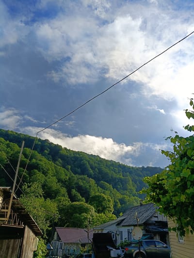 A medium shot captures a view of the city of Kepsha, Russia, showcasing a residential area nestled at the base of a verdant, tree-covered mountainside under a partly cloudy sky. In the foreground, wooden structures, including a house with a sloped roof and siding, are visible on the left. To the right, another house with a dark gray, corrugated roof and stone walls is situated, with a gray pickup truck parked in front of it. A satellite dish is mounted on the roof of this house. The middle ground is dominated by the lush green slopes of the mountain, densely populated with trees. A power line stretches diagonally across the frame from the top left to the top right. The sky above is a mix of deep blue with dramatic, dark gray storm clouds on the left, transitioning to brighter blue with white, fluffy clouds and patches of sunlight on the right. Green foliage from a vine is in the upper right corner. The overall scene suggests a rural or semi-rural setting, possibly experiencing changing weather.