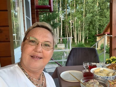 The image depicts a woman with short blonde hair and glasses, wearing a white t-shirt and a necklace, taking a selfie on a wooden porch overlooking a forest of birch trees. She is seated at a table with a meal, which includes a bowl of dumplings, a dish of fried food, and glasses of red liquid. The table is covered with a brown tablecloth, and a dark chair is visible behind her. The background shows the dense birch forest, characteristic of rural areas like Суроватиха, Russia. The lighting suggests it is late afternoon or early evening.