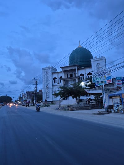 The image shows a street scene in Keudelapang, Indonesia, taken during twilight or dusk. A white multi-story building with a large, turquoise dome stands prominently on the left. Signboards for "BSI Link" and other businesses are visible on the buildings to the right of the mosque. Electrical wires crisscross the sky above. A paved road stretches into the distance with faint reflections from streetlights and vehicle headlights. Several motorcycles are on the road. The sky is blue with scattered clouds. A tree is growing in front of the mosque.
