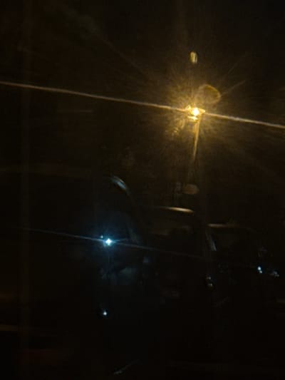 The image depicts a dark, nighttime scene, likely taken through a window, given the reflections and streaks of light. The primary source of light appears to be a streetlamp with a distinct yellow glow, casting rays across the dark environment. In the foreground, the silhouettes of vehicles are visible, their shapes obscured by the low light conditions. Reflections of what appear to be other lights, possibly from the streetlamp and vehicle headlights, are present on the surface through which the photo is taken. The overall impression is one of a dimly lit street or parking area in Stoevtsi, Bulgaria, at night.