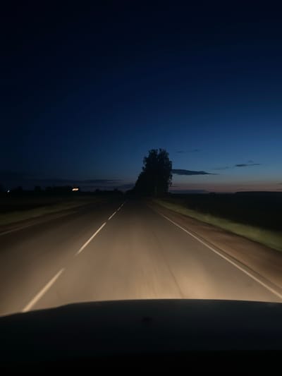 The image is a point-of-view shot from inside a vehicle driving on a paved road at dusk or night. The headlights illuminate the road ahead, which has white lines marking the lanes. The sky is a deep blue, transitioning to lighter hues near the horizon where the sun has set. Silhouettes of trees and possibly buildings are visible along the sides of the road. The location is specified as Aktsyabreski siel'ski savec, Belarus. The scene is rural and deserted, with no other vehicles or people visible. The overall impression is one of solitary travel through a quiet, darkening landscape.