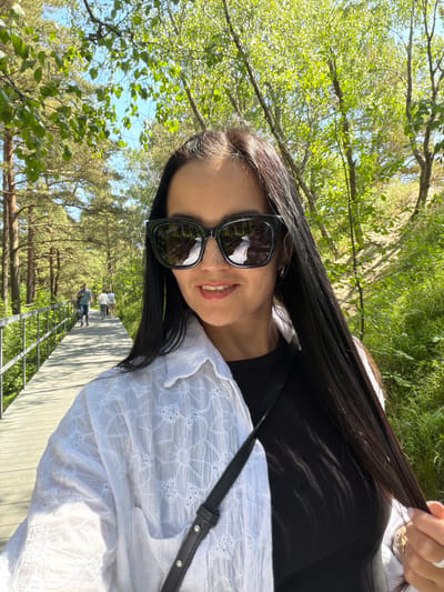 In the city of Morskoe, Russia, a woman with long, dark hair, wearing large black sunglasses and a white embroidered shirt over a black top, takes a selfie. She is walking on a wooden boardwalk, which curves to the left and is bordered by a metal railing. The boardwalk leads through a sunlit area with trees and lush green foliage on either side. In the background, two individuals are walking away on the boardwalk, and the sky is visible through the branches of the trees. The scene appears to be a park or a nature trail on a bright, sunny day.