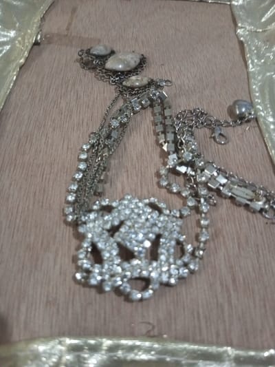 A silver-toned necklace or decorative chain is displayed on a light brown, wood-grain surface, likely indoors in Chandragiri, Nepal. The item is characterized by multiple interconnected chains and decorative elements. One end features three irregularly shaped, light brown and white variegated stones, each encased in a silver-toned metal setting, connected by small chains. From this section, several strands extend: some are simple thin chains, others are adorned with a continuous line of small, clear rhinestones, and some consist of flat, rectangular silver-toned links. A large, ornate central piece, heavily encrusted with clear rhinestones forming a complex, openwork floral or scroll pattern, is prominent. Towards the other end, a lobster clasp and an extender chain with a small spherical charm are visible, indicating it is an adjustable closure. Portions of a blurred, golden, shiny fabric are present along the left and bottom edges of the frame, bordering the wood surface. The item is static, showing no interaction.