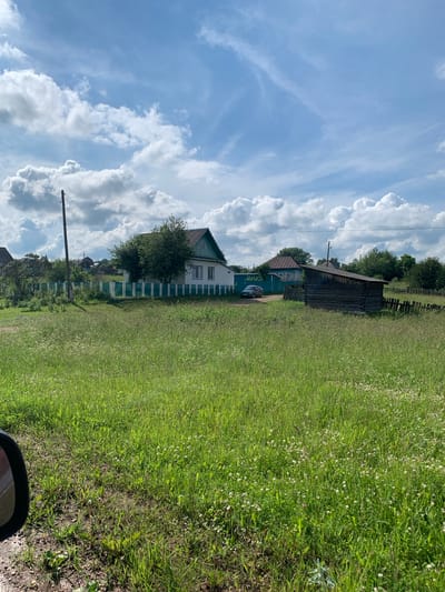 The image depicts a rural landscape under a partly cloudy sky. In the foreground, a vast field of green grass, dotted with small white wildflowers, stretches towards the background. A portion of a car's side mirror is visible on the far left, suggesting the photo was taken from a vehicle.
Further back, several wooden structures and houses are visible. A white house with a green roof and trim stands prominently, bordered by a green and white picket fence. Next to it, a dark, rustic wooden shed with a sloped roof is situated. Behind these buildings, more houses and trees suggest a small village or settlement. A single utility pole with electrical wires stands to the left of the white house. The environment appears to be a rural area, likely a village in the countryside. The time of day appears to be daytime, given the bright lighting and shadows. There are no people visible in the image.