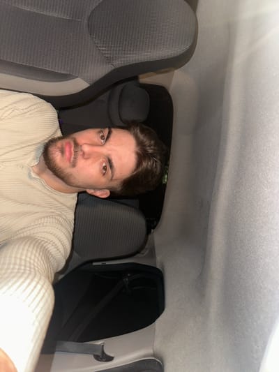 A medium-shot selfie shows a young man with brown hair and a beard inside a car. He wears a cream-colored, button-up shirt. The interior of the car consists of gray fabric seats and a white roof. The location and city of Rafz, Switzerland, cannot be determined from the provided image.