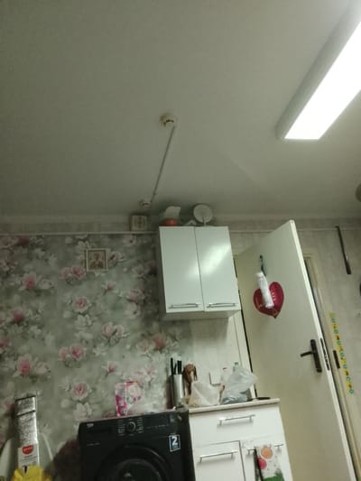 The image shows a cluttered kitchen area. On the left, a floral wallpaper covers the wall, with a small framed picture hanging on it. A silver-wrapped item, possibly a food product, is on the counter next to a black washing machine. Above the washing machine, there is a pink and white box. To the right of the washing machine, a white countertop unit with drawers is visible, holding various items including plastic bags, bottles, and utensils. A white cabinet with two doors is mounted above the countertop. On the door of a white cupboard, a red heart-shaped decoration is hanging with a white cylinder. A white door is on the far right of the image, with a black handle and a strip of colorful decorative tape running vertically alongside it. The ceiling has a bright fluorescent light fixture. There are no people visible in the image. The overall impression is of a lived-in and functional kitchen space.