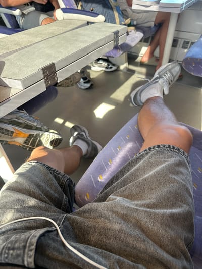 This image shows a person's legs and feet sitting on a train. The person is wearing gray denim shorts and white socks with gray sneakers. The train seats are upholstered in a purple fabric with a subtle yellow pattern. A folding table is visible in the foreground, attached to the seat back in front. The train appears to be in motion, as indicated by the sunlight streaming through the windows and casting shadows. The background shows other passengers seated on the train. The image was taken on a train traveling between Perpignan and Salses-le-Château in France.