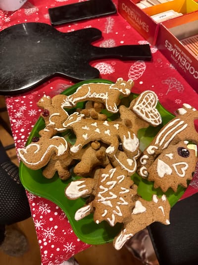 A festive scene shows a green plate piled high with gingerbread cookies of various shapes: bears, hearts, hedgehogs, a dragon, and other animals. The cookies are decorated with white icing. The plate sits on a red tablecloth with a white snowflake pattern, next to a box of Codenames. A black guitar-shaped object and a black phone are nearby. The image has a warm, inviting feel.