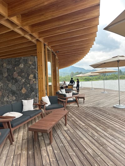 The image shows an outdoor seating area on a wooden deck overlooking a body of water. The deck is furnished with dark gray sofas and brown wooden coffee tables. In the background, there are several beige umbrellas providing shade. Beyond the deck, a calm body of water is visible, with lush green hills and mountains in the distance under a cloudy sky. Two individuals, one in a black shirt and the other in a black top and pants, are standing near a counter or bar area. Another person, wearing a red cap, is seated at a table in the far left background. The architecture of the building features a wooden slatted roof and a stone wall. The overall scene suggests a serene and upscale lakeside or waterfront establishment. The image does not provide any specific visual cues to indicate it is located in Santa Teresa Tiloxtoc, Mexico.