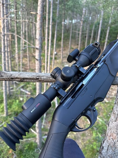 A black semi-automatic rifle with a scope is resting on a wooden branch in a forest. The rifle is positioned diagonally, with the scope on the left and the stock on the right. The scope is black and has a rubber eyepiece. The rifle has a black grip and a black trigger. The forest appears to be made up of tall, slender trees with a light brown bark. The ground is covered in green foliage. The lighting suggests it is daytime. There are no people visible in the image. The scene is likely in a rural or natural setting.