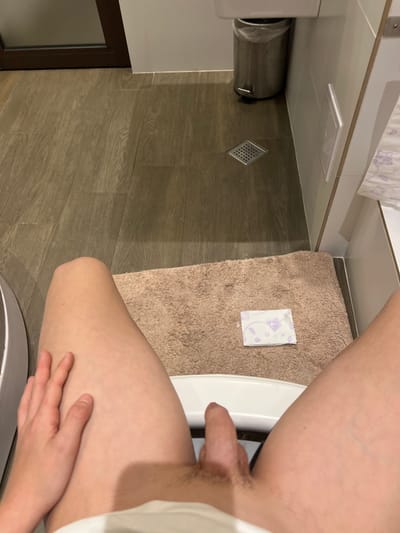 A person is standing in a bathroom with their penis exposed. The bathroom has a tiled floor, a toilet, a bathmat, and a sanitary napkin wrapper on the floor. The location is not specified.