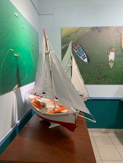 The image shows a wooden table with two sailboat models on it. Behind the models, there are two large wall-mounted photographs of boats in water. The first photograph on the left depicts a large expanse of bright green water with several small white boats, some with nets, and faint shadows cast by the boats on the water. The second photograph on the right shows an aerial view of boats in greenish-blue water near a shoreline. One boat is blue and white, another is primarily white with some colored accents, and a third is mostly white with a wooden deck and people visible. Several smaller objects, possibly debris or other boats, are scattered in the water. The sailboat models in the foreground are white with orange and red accents. The model in the front is detailed with rigging, sails, a mast, a ladder, and an anchor. The second model behind it is partially obscured by the first. The room appears to be indoors, with a white wall behind the photographs. The table is made of polished wood. The location is identified as Boca del Río, Venezuela.