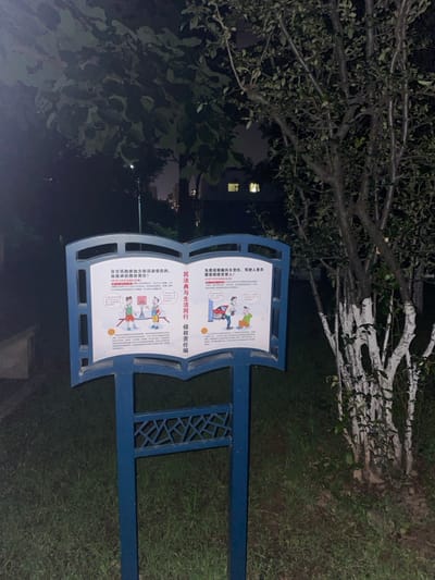The image depicts a blue, book-shaped public information sign positioned outdoors at night in a park or residential area of Dali City, China. The sign stands on a metal base with an ornate, geometric pattern. Its two white panels display cartoon illustrations and Chinese text, educating the public on civil law, specifically the "Civil Code and Life Go Hand in Hand: Tort Liability Section."

The left panel illustrates three cartoon figures on a basketball court: one in red/white mid-jump with a ball, another in green observing, and a third in blue collapsed on the ground, implying an injury during recreational activity. The right panel shows two cartoon figures next to a red car on a hydraulic lift; one in red appears to be working on the car, while the other in green sits on the ground, seemingly injured, with a thought bubble above their head.

The environment is dark, with an overhead street light faintly visible in the upper left. Dense green foliage, including a tree with white-painted lower trunks and branches on the right, surrounds the sign. Overgrown grass and dirt are visible at the base of the sign. In the distant background, illuminated windows of multiple-story buildings suggest a residential or urban setting.
