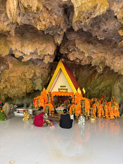 The image depicts the interior of Tham Phung Chang, a cave temple located in Phang-nga, Thailand. The cave's natural ceiling and walls are characterized by rugged rock formations and stalactites, illuminated by a warm, yellowish light. The cave floor is covered with polished, light-colored ceramic tiles, displaying reflections of the scene.

A central shrine with a golden and red-gabled roof, typical of traditional Thai architecture, is positioned at the rear of the cave. This shrine contains various smaller statues and is adorned with offerings. Numerous statues, predominantly depicting elephants or elephant-headed deities, are arranged around the main shrine, particularly to its right. These statues are heavily draped with vibrant orange marigold garlands. A golden elephant statue rests on the tiled floor to the left of the main shrine, while a silver-toned elephant statue is present on the right. In the far left background, a raised platform holds a collection of smaller seated figures, identified as Buddha statues or monks, alongside a large ceramic urn.

Two individuals are present, kneeling on the tiled floor, facing the central shrine in a posture of veneration. The individual on the left wears a red and dark-colored long-sleeved top with yellow accents and a face mask. The individual on the right wears a dark, long-sleeved garment. Both have their hair tied back.