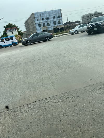 The image depicts a street scene in Igando Oloja, Nigeria, under an overcast sky. The foreground consists of a wide, light-colored concrete road surface with visible cracks and expansion joints. Multiple vehicles are present on the road, suggesting active traffic. On the far left, a blue and white minivan is positioned near a white building with a red triangular roof and a sign displaying "PEACE GARDEN." Adjacent to this, a dark grey sedan is central in the mid-ground, followed by a silver sedan further right. A black Lexus SUV with a visible Nigerian license plate (LAA 34JL) occupies the far right of the frame. In the background, a multi-story building under active construction is prominent, clad in blue sheeting and scaffolding. Another partially constructed concrete building is visible to its right. Overhead power lines traverse the scene in the upper-mid section. No discernible individuals are clearly visible.