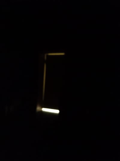 A dimly lit scene in Жаңатұрмыс, Kazakhstan, features a rectangular frame composed of a vertical yellow line on the left, extending upwards and then horizontally to the right, with a bright, blurry horizontal white light at the bottom left. The overall environment is dark, suggesting a nighttime or indoor setting with minimal illumination.