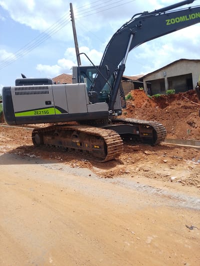 This media file shows a Zoomlion ZE215G excavator at a construction site in Iseyin, Nigeria. The excavator is the primary object, with its tracks and a portion of its boom visible. It is positioned on a dirt and gravel surface, with piles of reddish-brown soil to the right. In the background, there are some buildings with corrugated metal roofs and concrete structures. Power lines stretch across the sky, supported by a wooden utility pole. The sky is partly cloudy, suggesting daytime weather. The scene appears to be a typical construction or road work environment. There is visible text on the excavator's boom, reading "ZOMLION", and on its side, "ZE215G". The overall impression is one of ongoing infrastructure development.