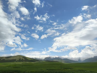 This media file depicts a scenic landscape of rolling green hills and mountains under a vast, cloudy blue sky. The foreground is dominated by a lush, vibrant green field, with some patches of yellow flowers visible towards the bottom. In the middle ground, gentle slopes rise, some covered in dark green trees forming a distinct pattern, while others are a lighter green, suggesting cultivated fields or natural growth. A lone, dark wooden pole stands in the middle of the field on the right side of the frame.
In the background, a range of mountains stretches across the horizon, partially obscured by dramatic, white clouds that are also scattered across the bright blue sky. The clouds are varied in size and texture, with some appearing wispy and thin, while others are thick and cumulus-like, casting shadows on the landscape below. The light suggests it is daytime, likely mid-morning or afternoon, given the brightness and the presence of clouds. The overall mood is peaceful and serene, conveying a sense of open space and natural beauty. There is no visible text in the image. The location context provided is Alagyaz, Armenia, which aligns with the mountainous and hilly terrain.