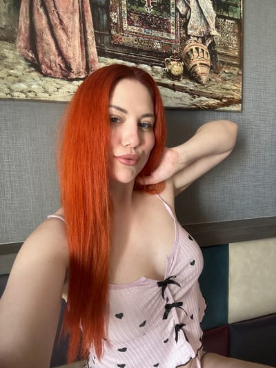 In a selfie taken indoors, a woman with long, bright red hair is positioned in the foreground. She is wearing a light pink camisole adorned with black heart patterns and black ribbon ties. Her left arm is bent and resting behind her head, while her right hand is near her neck. Her gaze is directed towards the camera, and her lips are slightly parted. Behind her, a painting hangs on a textured grey wall. The painting depicts draped fabric and pottery. The lower portion of the image shows the back of a sofa with alternating colored sections. The overall setting does not contain any identifiable landmarks or clues to suggest a specific location in Avcılar, Turkey.