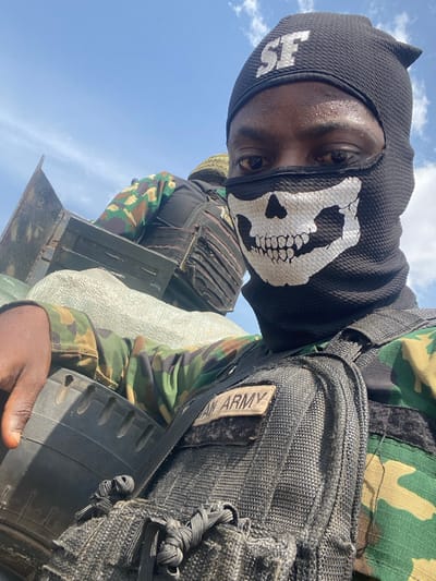Two individuals in military attire are captured in a daytime scene, likely within a vehicle, under a clear blue sky with scattered clouds. The foreground individual, dark-skinned, is taking a selfie. This person wears a black balaclava featuring a white skull design over the lower face and "SF" in white text on the forehead. Visible are dark eyes, and sweat on the exposed forehead. A camouflage uniform is worn beneath a black tactical vest, which bears a "NIGERIAN ARMY" patch. The right hand rests on a black, ribbed object, resembling a tire. A second individual is partially visible in the background, to the left, also in camouflage attire and a dark tactical vest. Surrounding objects include light-colored, bulky sacks and dark, rectangular containers, possibly ammunition boxes, along with a metal vehicle panel. This scene is located in the city of Gburuku, Nigeria.