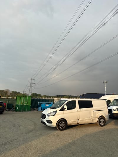 The image, oriented horizontally, captures an outdoor industrial or commercial yard under an overcast sky in Mid Suffolk, United Kingdom. The foreground shows an asphalt or gravel surface. Two white commercial vans, possibly Ford Transit Customs, are prominently parked. The closest van has the visible license plate "NG67 RDO". Another white van behind it features an "EU" sticker on its front grille. Further back, stacked green shipping containers form a boundary, behind which a line of trees is visible. In the mid-ground, a tall metallic lattice-work electricity pylon stands against the sky, with multiple power lines stretching diagonally across the frame. A second pylon is faintly visible in the far distance. A lamp post equipped with floodlights is also present near the vans. The sky is uniformly grey and cloudy, indicating overcast weather. No specific activity or individuals are depicted. A small black car with the license plate "MA22 CVW" is partially visible in the bottom left corner. The overall scene suggests a quiet, possibly late afternoon or early evening setting, given the soft, diffused light.